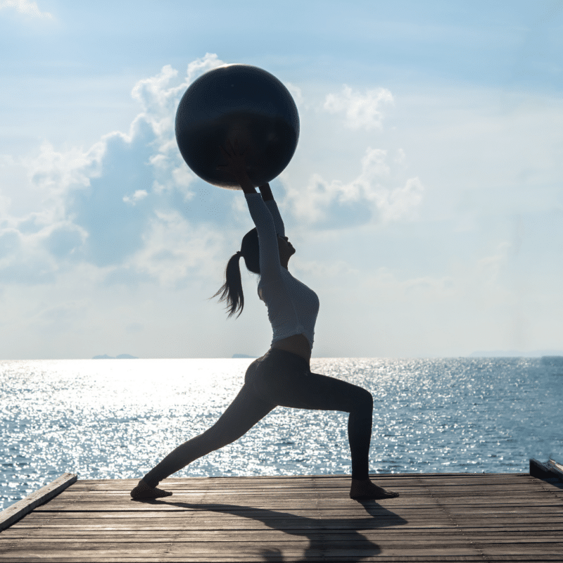 Kvinde laver øvelse med fitnessbold på en mole ved havet