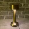 En stilfuld, gylden bordlampe med en cylindrisk base og top, oplyst foran en murstensvæg.
