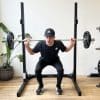 En person iført sort træningstøj og kasket laver squat med vægtstang på en træningsstation. I baggrunden er der en grøn plante ved en hvid væg.