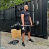 En mand står udendørs foran en sort container, iført sort træningstøj. Han løfter en tung vægtstang med flere skiver i et træningsområde. Rundt om er der træningsudstyr, herunder en træboks og ekstra vægtskiver.