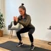 En kvinde i træningstøj udfører en squat-øvelse med en elastik om anklerne på en sort træningsmåtte i et lyst rum. I baggrunden ses en stol, en plante og et lille bord med en lampe.