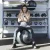 Kvinde sidder på en TRX-bold i et fitnesscenter og kigger på sin telefon.