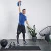 En mand i blå træningsskjorte og sorte shorts løfter en blå kettlebell over hovedet. Han står på en træningsmåtte i et minimalistisk rum med en hvid væg, en plante i en grå potte, en stol, og forskellige træningsudstyr som en vægtstang og medicinbold ved siden af ham.