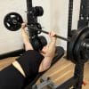 Kvinde udfører bænkpres på en træningsbænk i et fitnesscenter. Hun ligger på ryggen og løfter en vægtstang med skiver. Vægtstativ og flere vægtskiver ses i baggrunden.